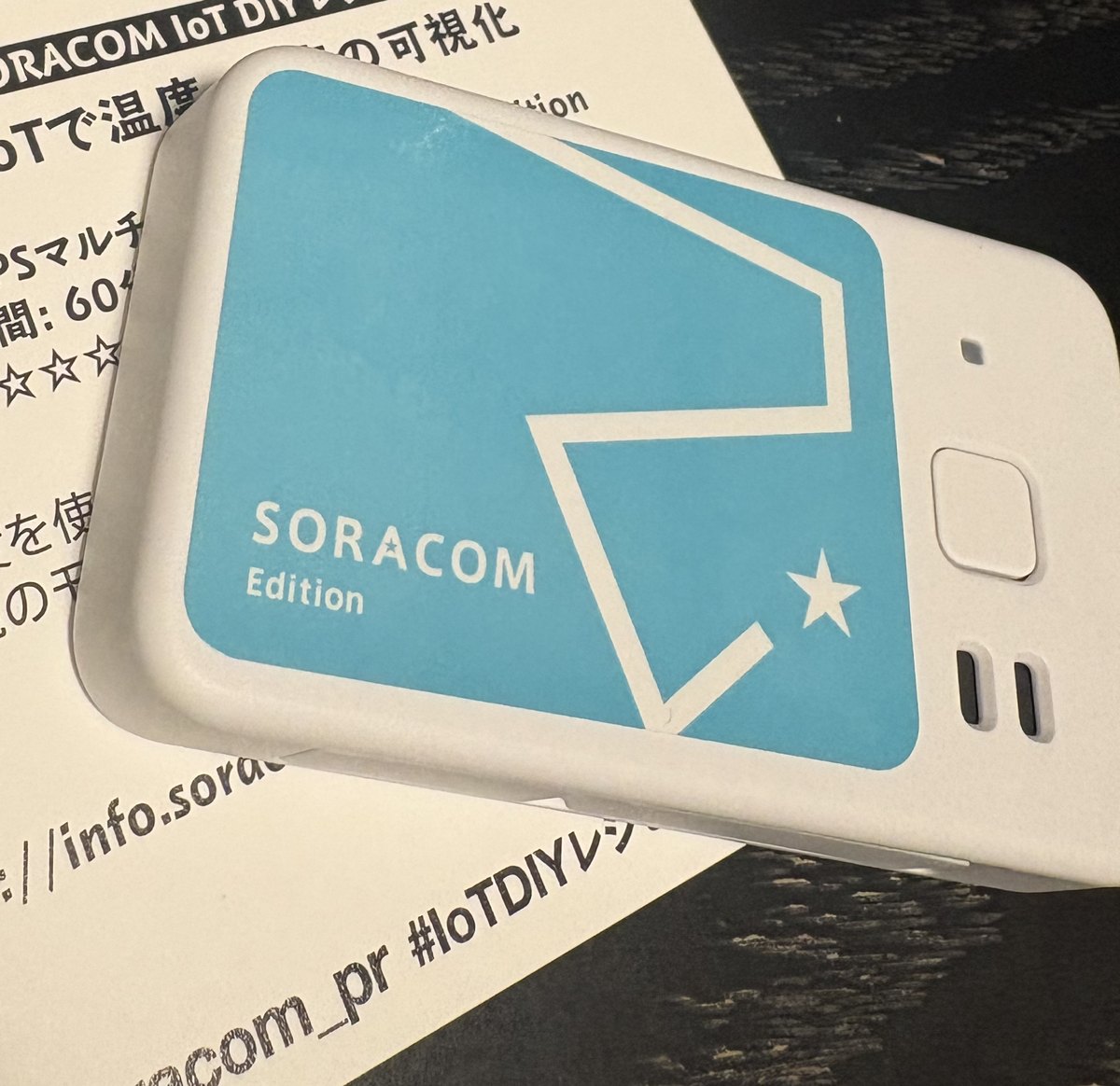 GPS マルチユニット SORACOM Edition の本体。白い樹脂のケースに水色の SORACOM ロゴのシールが貼られ、側面に充電端子とボタンがある。下には SORACOM の IoT DIY レシピの紙が敷いてある。